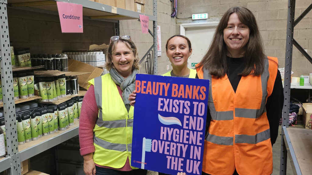 3 women stand in a warehouse with a banner that shows Beauty Banks