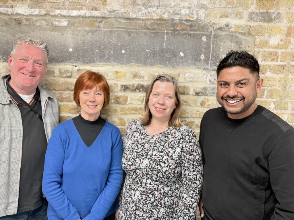 Four people stand in front of a brick wall. They are smiling.