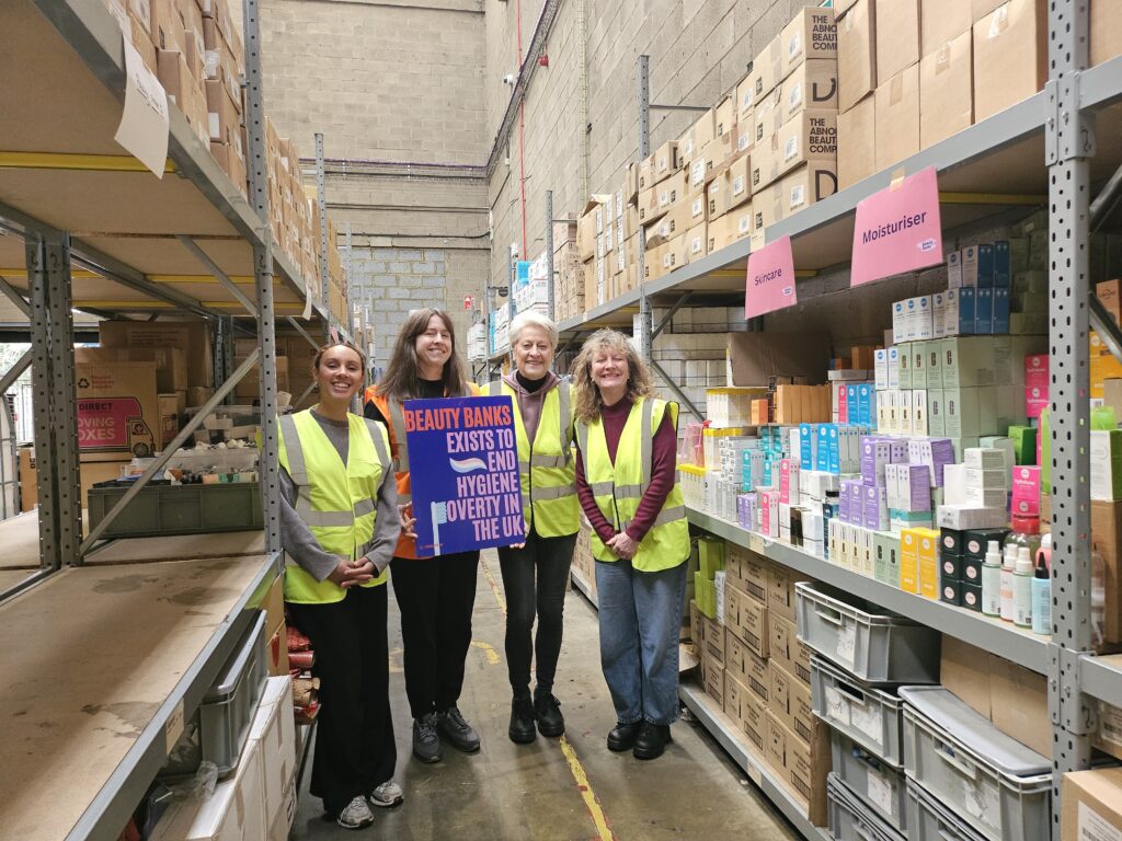 4 women stand in a warehouse with a banner that shows Beauty Banks