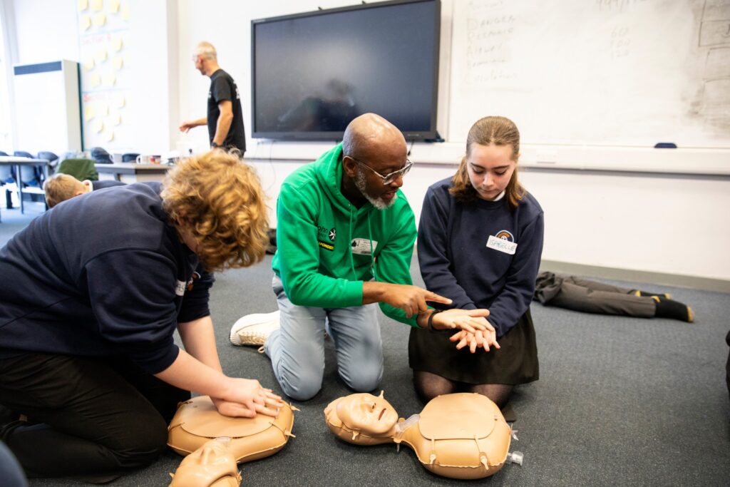 An adult in a green top shows two young responders how to perform cpr.