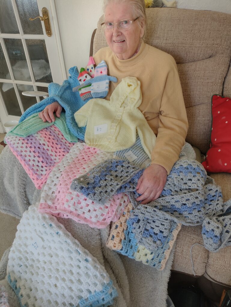 A lady sits in a chair covered in knitted items.