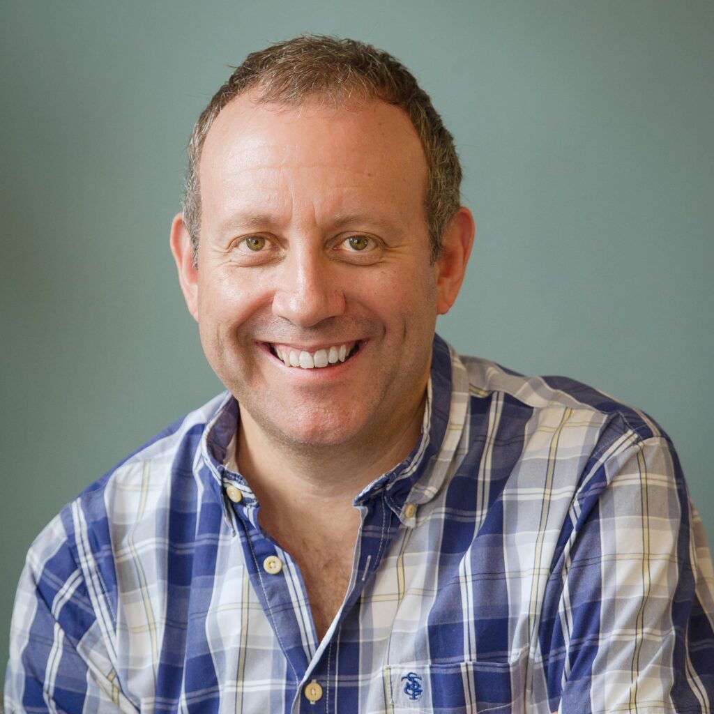 A man is smiling. He wears a checked shirt